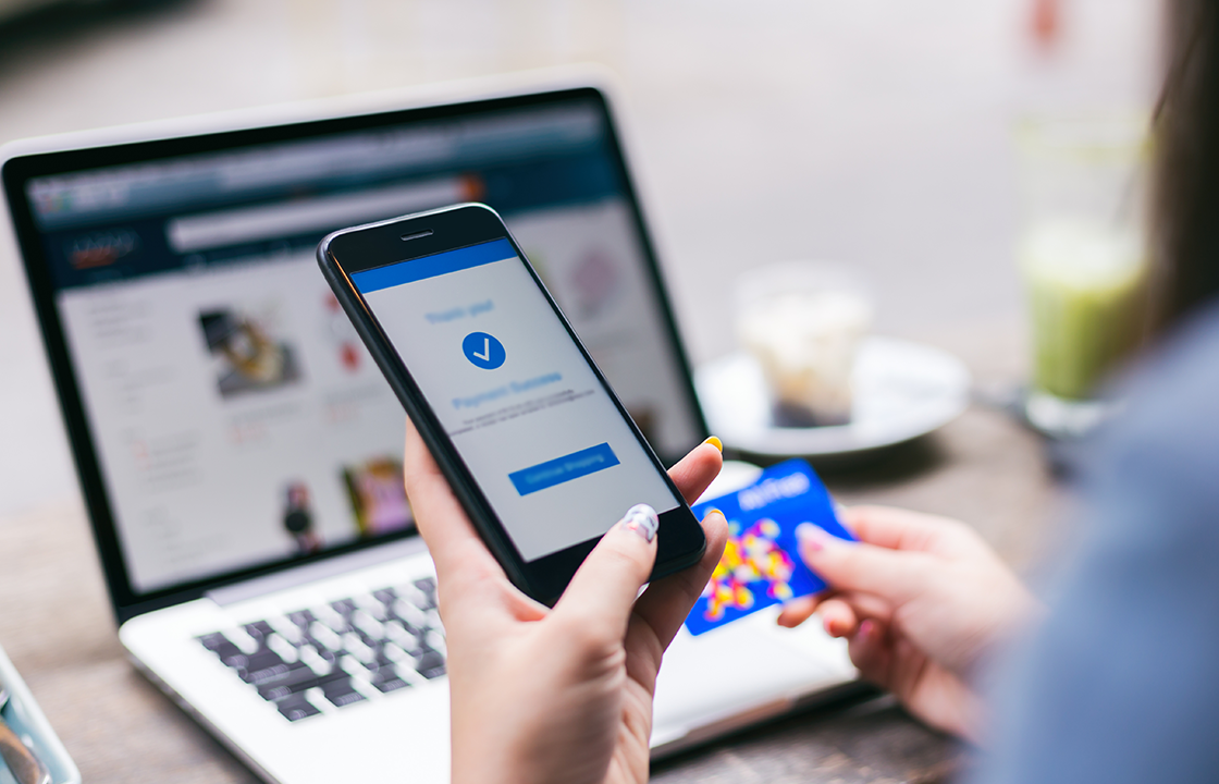 Téléphone portable à la main pour effectuer un achat en ligne, avec un ordinateur portable en arrière-plan et une femme tenant une carte de crédit